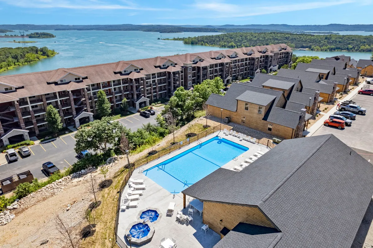 A large outdoor swimming pool and hot tub area sit beside lush greenery and a lake in the background. Modern apartment buildings are visible, and the spacious area is designed for sunbathing, swimming, and socializing.