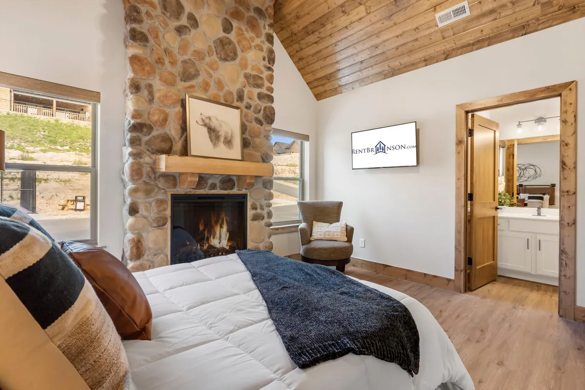 A warmly lit bedroom featuring a plush bed with decorative pillows, a prominent stone fireplace, large windows offering scenic views, and a rustic wooden door. The ambiance is cozy and inviting, with the fireplace serving as the room’s focal point and natural light streaming in through the windows.