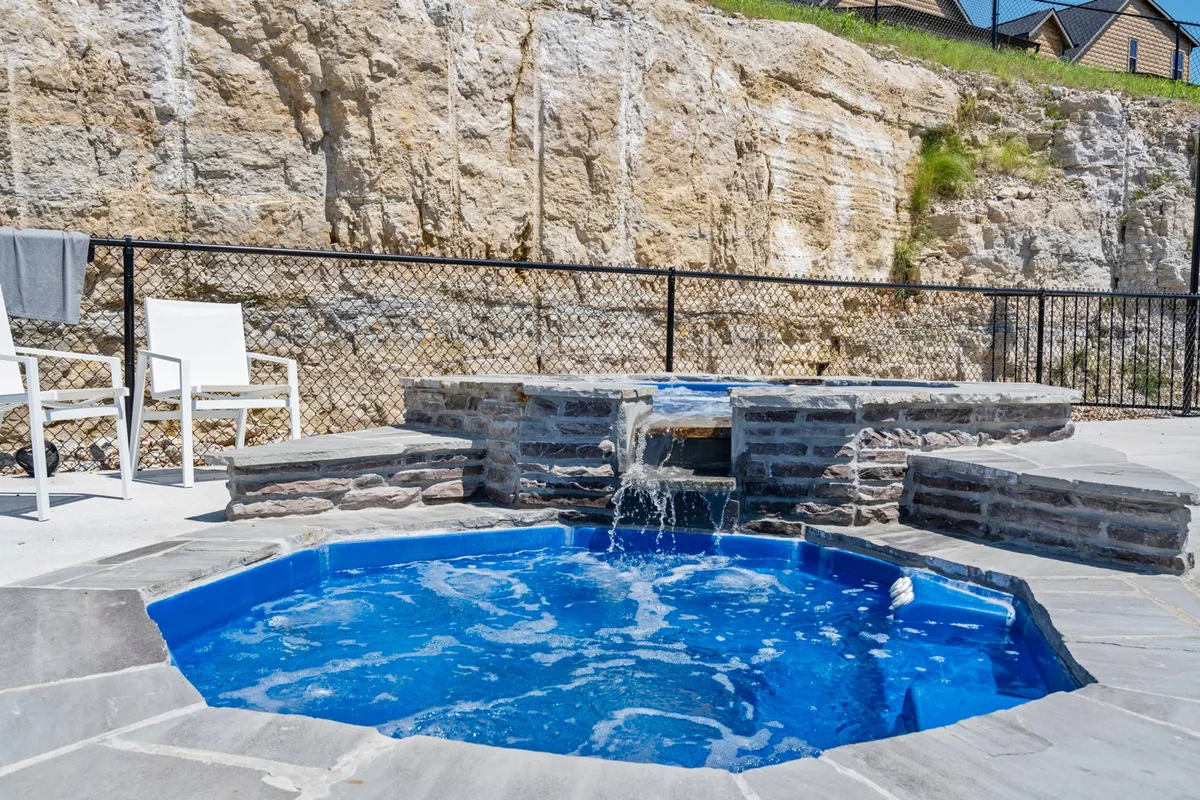 A private hot tub is the focal point, set amidst a rocky landscape with lounge chairs nearby. The space feels secluded and tranquil, enhanced by the soft glow of sunset and hints of water views in the background. Friends are shown relaxing, suggesting a social yet peaceful atmosphere.