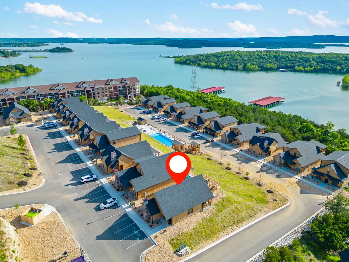 A wide exterior view shows a sparkling outdoor swimming pool in the foreground with lounge chairs, a clear lake just beyond, and a row of inviting log cabins. The scene also includes a parking area and is framed by trees and greenery, conveying a resort-style environment.