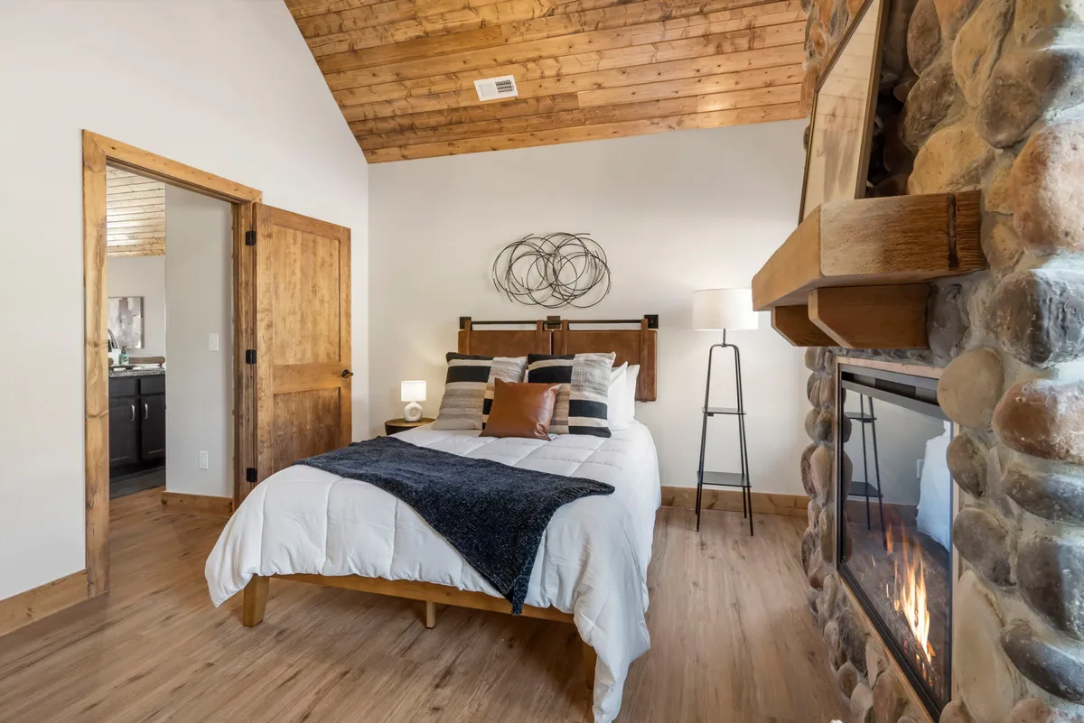 A comfortable bedroom with a large bed adorned with decorative pillows, set against a backdrop of a stone wall and wooden ceiling. A fireplace sits nearby, adding warmth and charm to the space, while soft lighting enhances the sense of intimacy and tranquility.