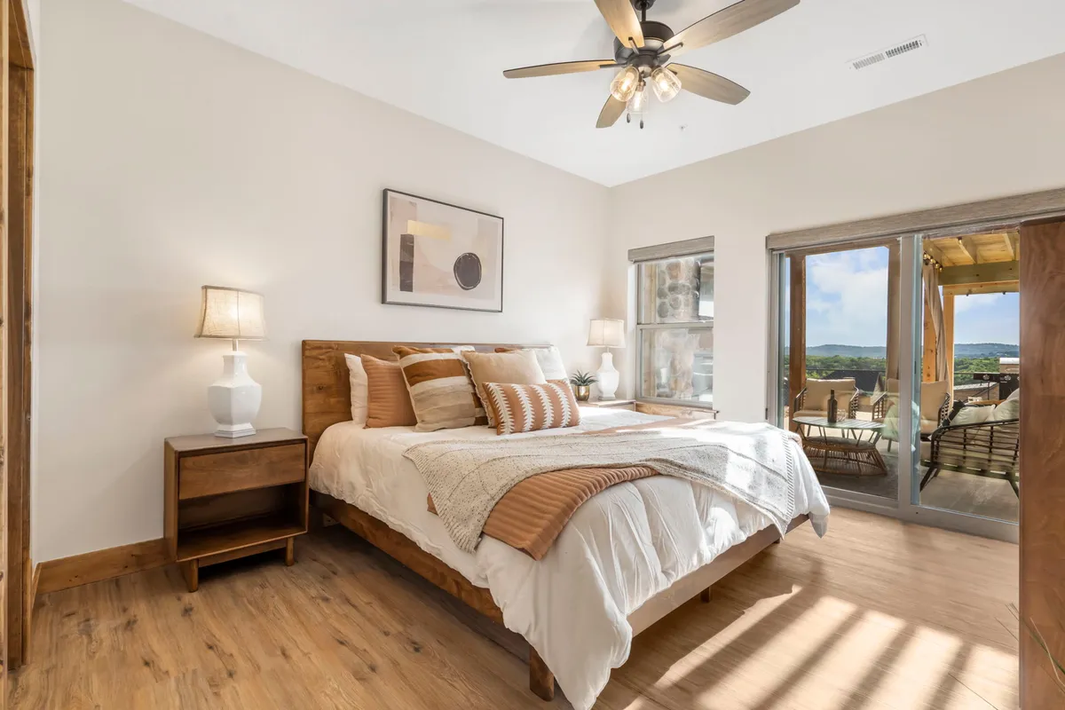 A sunlit bedroom with a large bed dressed in decorative pillows, a nightstand with a lamp, and a wide window revealing scenic views. The room feels cozy and restful, inviting guests to enjoy morning coffee in bed or unwind in peaceful seclusion.