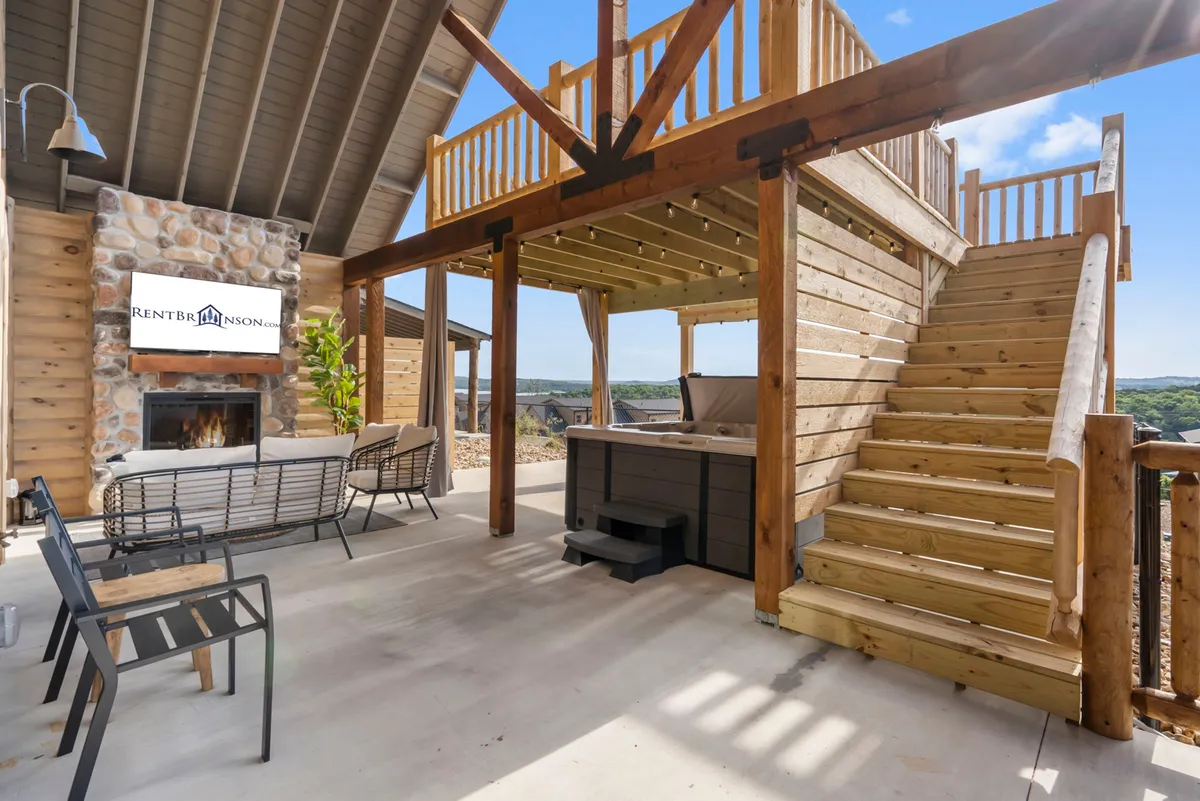 The image showcases the exterior of a charming log cabin featuring a cozy outdoor seating area, a bubbling private hot tub, an inviting stone fireplace, and a staircase leading to an upper deck perfect for stargazing. Surrounded by the natural beauty of the Ozarks, the setting is ideal for relaxing, socializing, and enjoying scenic views.