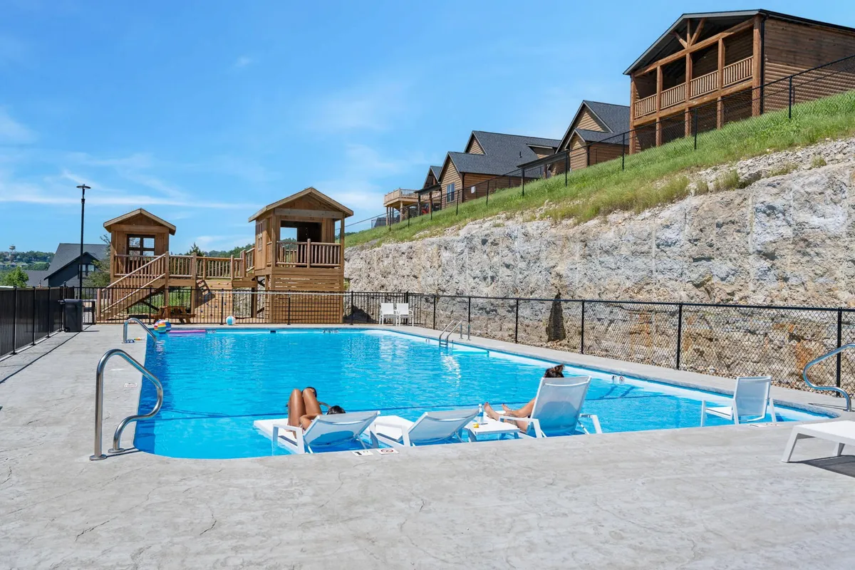 An outdoor pool scene featuring sparkling water, wooden play structures for children, and a row of lounge chairs inviting sunbathers. The setting is bright and relaxed, with children playing nearby and plenty of space for families to gather and unwind together.