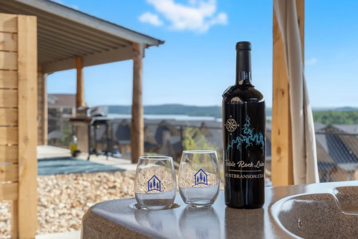 A cozy outdoor table is set with a bottle of wine and two glasses, perfectly positioned to take in expansive views of the sparkling lake beyond. The setting is intimate, with an emphasis on relaxation and celebration amid natural beauty.